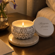 A lit scented candle in a white ceramic pot with an ornate gray pattern sits on a round marble coaster on a wooden bedside table. The white lid, printed with the brand name 'Thompson Ferrier', rests beside it.